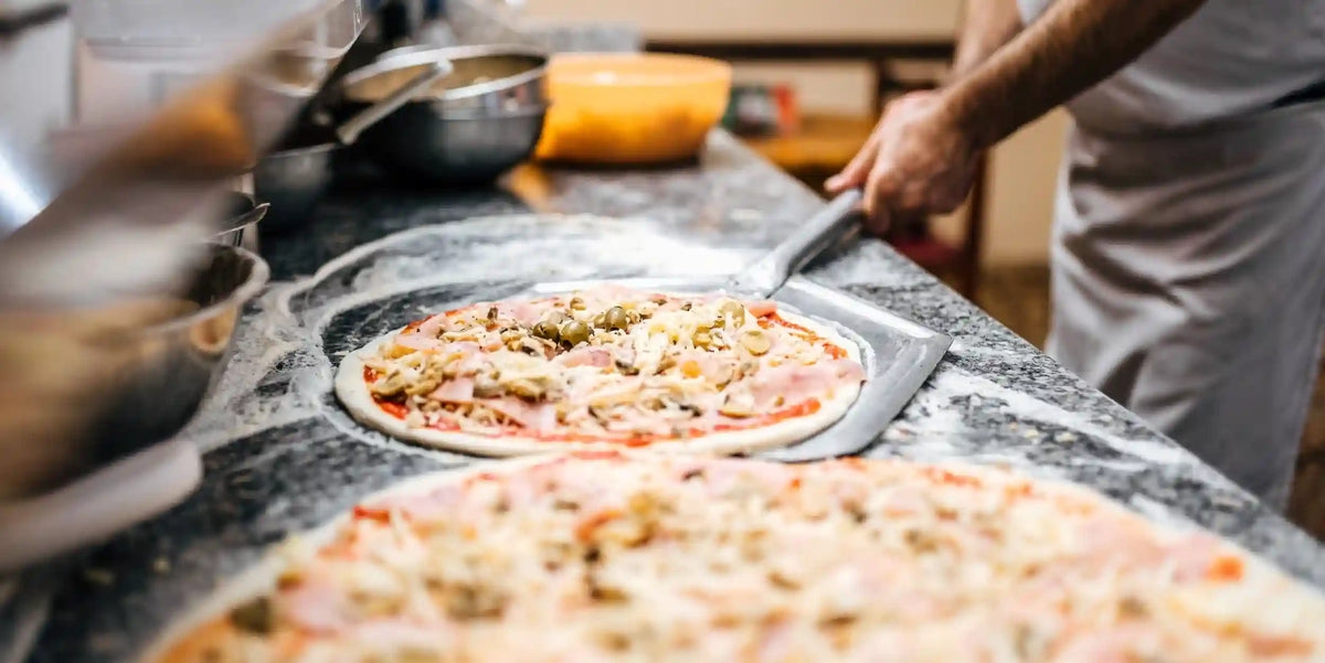Pizzabagare lägger en pizza med skinka, champinjoner och oliver på en pizzaspade på en mjölad stenbänk