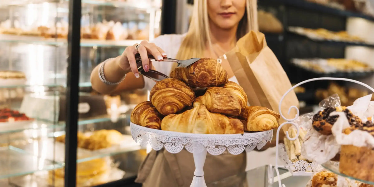 Personal plockar en croissant med tång från ett tårtfat och lägger den i en papperspåse, med bakverk i glasmontrar bakom
